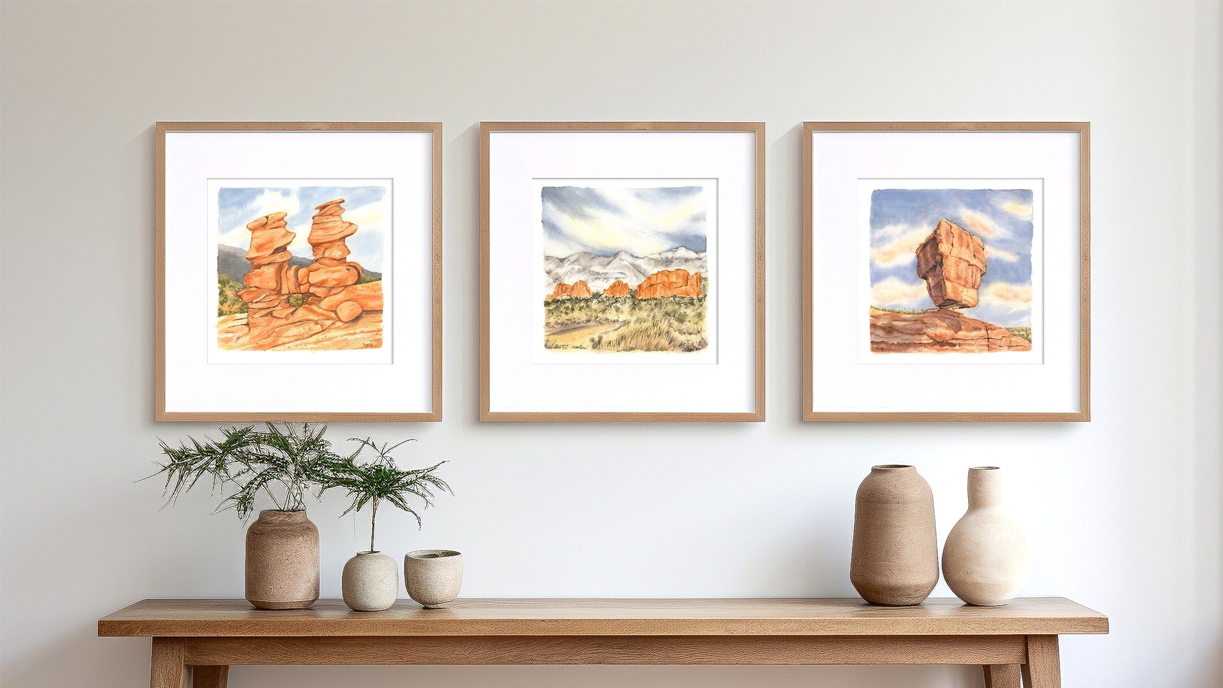 Three framed artworks of natural landscapes above a wooden bench with decorative vases and plants.