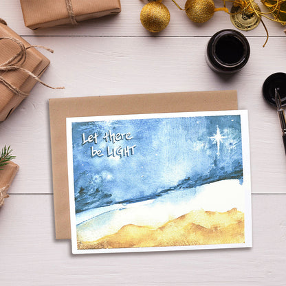 A greeting card with a watercolor painting depicting an abstract landscape with the text 'Let there be light!' The card is displayed alongside a envelope, decorative items, and a cup of tea.